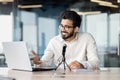 A smiling Indian young man is sitting in the office at a desk and talking through a microphone on a video call on a Royalty Free Stock Photo