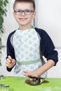 Smiling jagged boy in glasses and a kitchen apron squeezes shapes from metal molds in a brown batter.