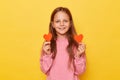 Smiling joyful positive optimistic little girl wearing pink sweatshirt holding red little hearts isolated over yellow background