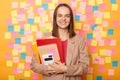 Smiling joyful woman teacher with brown hair wearing beige jacket holding paper folder, posing isolated over yellow background, Royalty Free Stock Photo