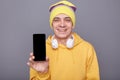 Smiling kind friendly man in beanie hat and yellow casual hoodie, holding mobile phone with blank screen, standing isolated over
