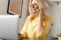 Smiling kind senior woman works from home. She speaks on the phone while sitting at the table In front of the laptop