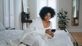 Smiling lady messaging smartphone at bedroom. Curly woman texting sms laying bed Royalty Free Stock Photo