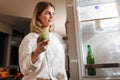 Smiling lady standing on kitchen at night and looking in open fridge holding green apple in hand Royalty Free Stock Photo