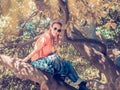 Smiling lady with two pigtails and a shirt tied on a belt sits on a tree in the park