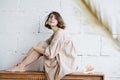 Smiling languid young woman in beige dress sitting on a low cupboard