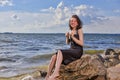 Smiling Laughing Winsome Relaxing Caucasian Brunette Girl Posing in Black Dress On Stone Line At Sea During Sunny Day Outdoors