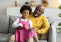 Smiling little african american girl sitting on knees of elderly grandfather on couch in living room interior Royalty Free Stock Photo