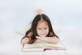 Smiling little Asian child girl with hardcover book lying on bed and looking camera over white background Royalty Free Stock Photo