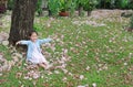 Smiling little Asian child girl sitting on green grass under tree trunk with falling pink flower in the park garden Royalty Free Stock Photo