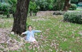 Smiling little Asian child girl sitting on green grass under tree trunk with falling pink flower in the park garden Royalty Free Stock Photo