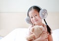 Smiling little Asian child girl wearing winter earmuffs and embracing teddy bear while sitting on the bed at home Royalty Free Stock Photo