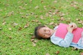 Smiling little Asian girl with book lying on green grass with dried leaves in the summer garden Royalty Free Stock Photo