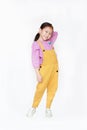 Smiling little Asian kid girl in pink-yellow dungarees poses touched hair keep back isolated on white background. Full length of Royalty Free Stock Photo