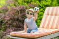 Smiling little baby boy is sitting on the deck chair and showing little soccer balloon Royalty Free Stock Photo