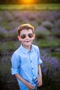 Smiling little boy standing on the lavender field. Funny child in sunglasses dressed in a blue shirt and a straw hat standing betw