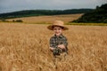 A smiling little farmer boy in a plaid shirt and straw hat poses for a photo in a wheat field. Heir of farmers