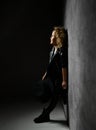 Girl with blond curly hair in black leather suit standing in spotlight close to dark wall looking up and holding hat in hand