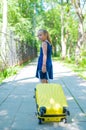 A smiling little girl in a blue dress is walking along the street with a yellow suitcase. A child is going on a summer