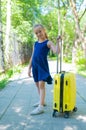 A smiling little girl in a blue dress is walking along the street with a yellow suitcase. A child is going on a summer