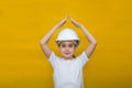 Smiling little girl in a construction white helmet holding hands above his head house, roof on a yellow background Royalty Free Stock Photo