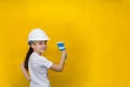 Smiling little girl in a construction white helmet holds a paint brush, standing back on a yellow background