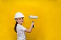 Smiling little girl in a construction white helmet holds a paint roller, standing back on a yellow background