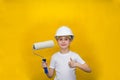 Smiling little girl in a construction white helmet holds a paint roller on a yellow background. concept of renovation