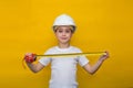 Smiling little girl in a construction white helmet with a construction tape measure in her hands on a yellow background