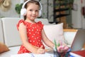 Smiling little girl in headphones handwrite study online using laptop at home