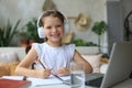 Smiling little girl in headphones handwrite study online using laptop at home