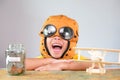 Smiling little girl in a pilot hat with coins in a clear jar for savings for travel and a wooden toy plane on the table. Childhood