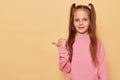 Smiling little girl with ponytails wearing pink sweatshirt posing isolated over beige background pointing away at copy space for Royalty Free Stock Photo