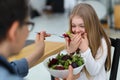 Smiling little girl refuses to eat raw salad, mother feeds her child with healthy food