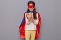 Smiling little girl wearing superhero costume and mask isolated over gray background using mobile phone for scrolling online Royalty Free Stock Photo