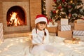 Smiling little girl wearing white sweater and santa claus hat, sitting on floor near Christmas tree, present boxes and fireplace, Royalty Free Stock Photo