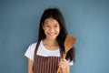 Smiling long brunette hair 10s Asian girl in brown stripped apron holding wood spatula for home cooking food with blue background