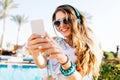 Smiling long-haired girl in sunglasses and earphones making photo of beautiful landscape with exotic palm trees. Joyful Royalty Free Stock Photo