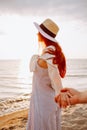 Smiling long haired woman in a hat holding boyfriends hand along empty ocean beach sand at sunset . Follow me concept