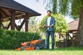 Smiling male landscaper in denim shirt trimming overgrown lawn with lawn mover at summer day.