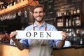 Smiling male owner of coffee shop is standing indoors. He is holding placard and inviting everybody to come in. Open Royalty Free Stock Photo