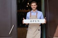 Smiling male owner of coffee shop is standing outdoors. He is holding placard and inviting everybody to come in. Open Royalty Free Stock Photo