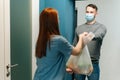 Smiling male wearing protective face mask delivering groceries bag to female housewife customer on doorway at home. Royalty Free Stock Photo