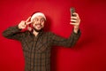 smiling man in Christmas hat takes selfie, takes pictures of himself on phone with hand showing victory peace sign Royalty Free Stock Photo