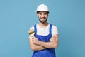 Smiling man in coveralls protective helmet hardhat hold paint brush isolated on blue wall background. Instruments Royalty Free Stock Photo