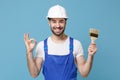 Smiling man in coveralls protective helmet hardhat hold paint brush isolated on blue wall background. Instruments Royalty Free Stock Photo