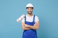 Smiling man in coveralls protective helmet hardhat hold paint roller isolated on blue background. Instruments Royalty Free Stock Photo