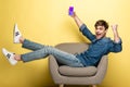 Smiling man in denim clothes holding smartphone with shopping app sitting on armchair on yellow