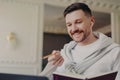 Smiling man freelancer in pullover and wireless earphones with pencil and book in hands having video call on laptop Royalty Free Stock Photo