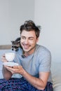 Smiling man with funny raccoon on shoulder holding coffee cup while sitting on bedding. Royalty Free Stock Photo